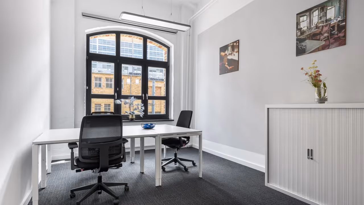 Zwei schwarze Bürostühle an weißem Tisch, großes Fenster, grauer Teppich, weiße Wände, Deckenleuchte, Kunst, Pflanzen.