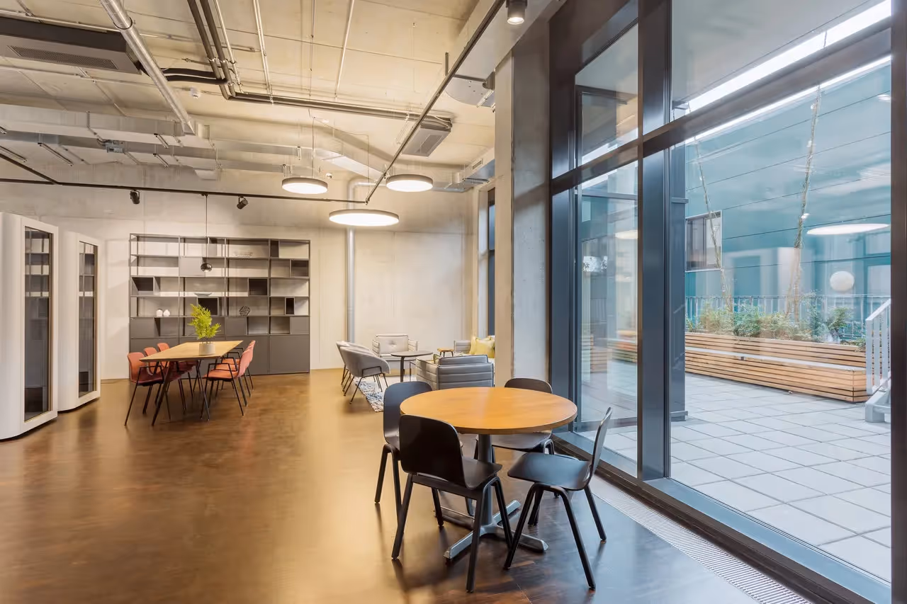 Runder Holztisch mit schwarzen Stühlen, große Fenster, Lounge-Bereich mit Sofas, Regale, Hängelampen, Holzboden.