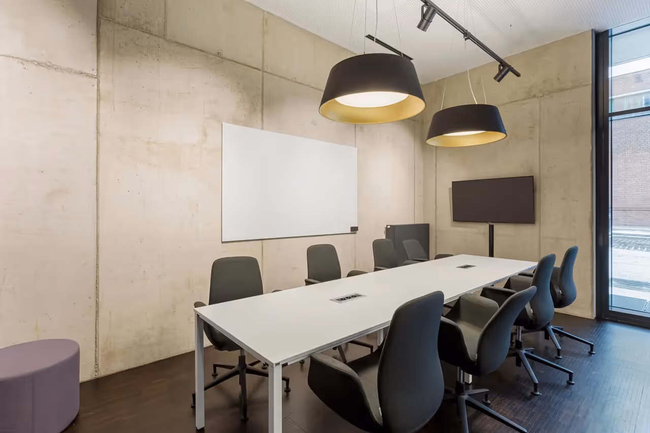Konferenzraum mit weißem Tisch, schwarzen Stühlen, Betonwänden, Hängelampen, Whiteboard und Bildschirm.