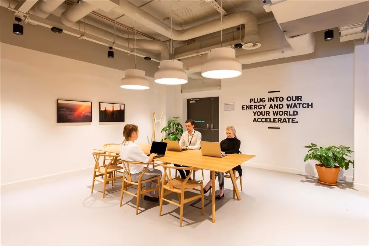 Drei Personen an Holztisch mit Laptops, helle Deckenleuchten, Pflanzen, Wandzitat, industrielle Decke, Bilder an Wand.