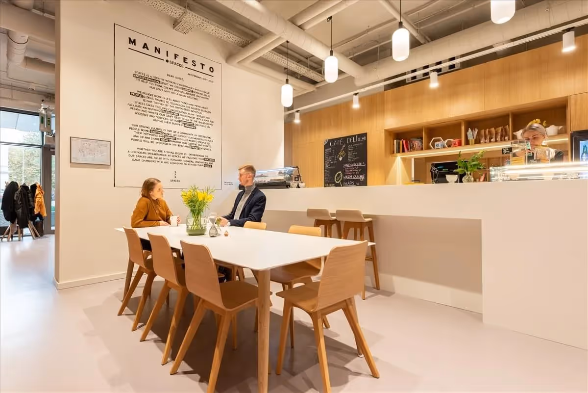 Heller Raum mit Holzmöbeln, zwei Personen am Tisch, Kaffetheke mit Barhockern, große Wand mit Manifest-Text.