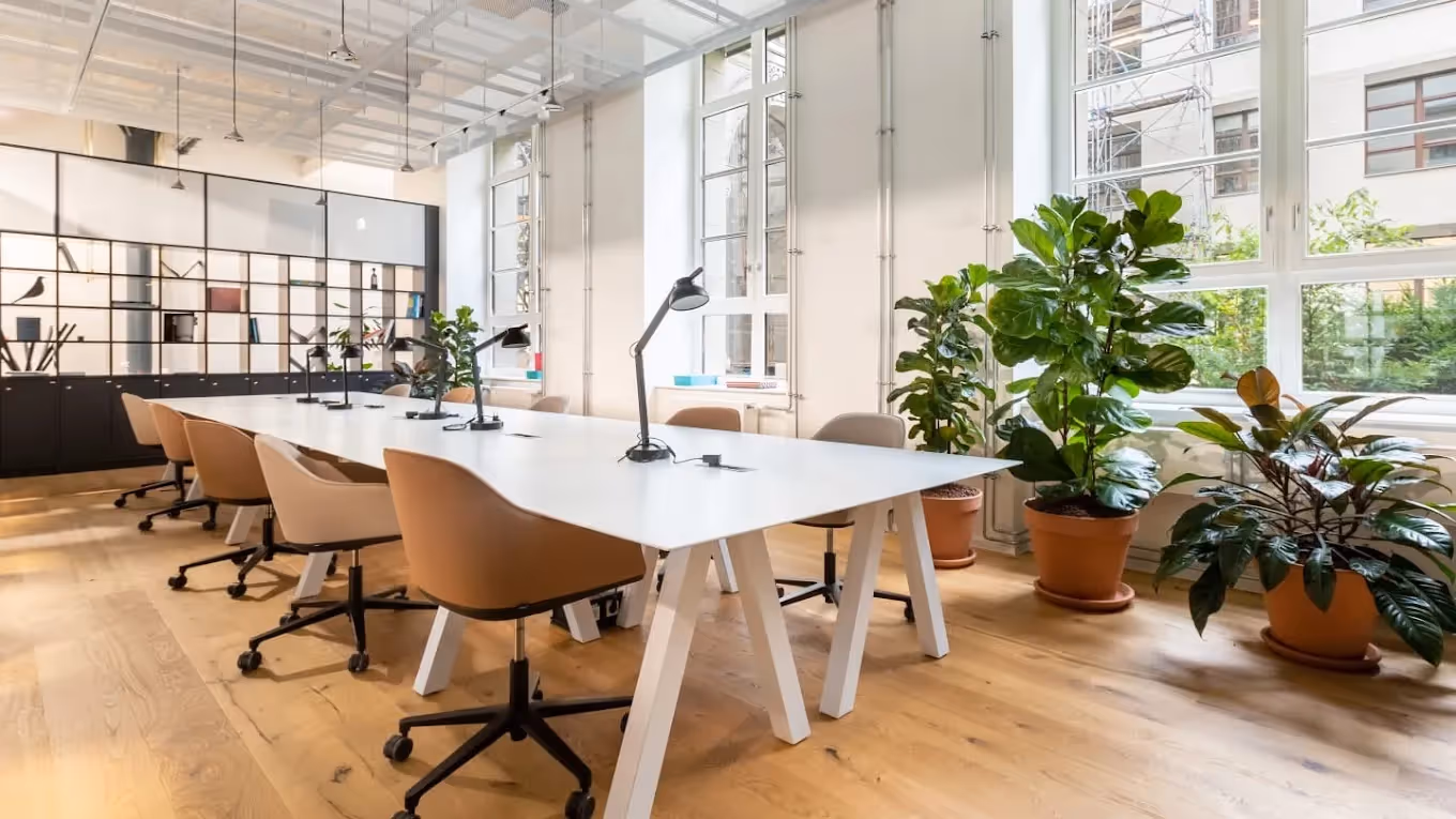 Heller Raum mit großem Tisch, braunen Stühlen, Pflanzen, Holzfußboden und großen Fenstern im Coworking-Space.
