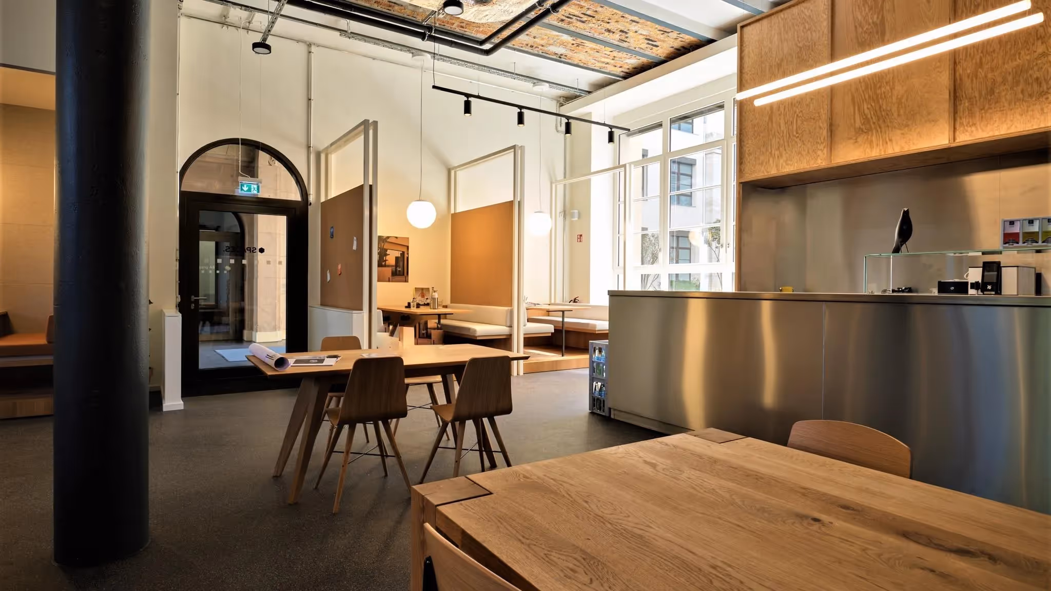 Helle Arbeitsbereiche mit Holztischen, Sitznischen, hohen Fenstern, Metalltheke und modernen Leuchten.