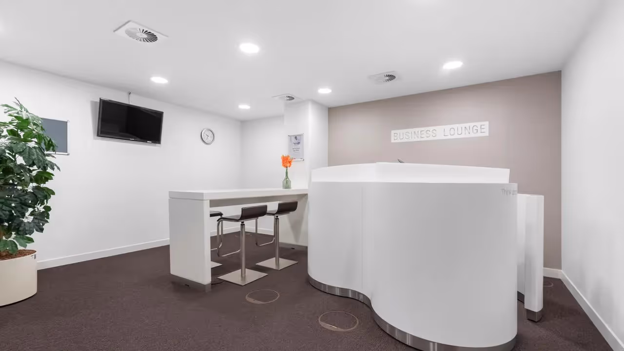 Heller Raum mit Tresen, Barhockern, Topfpflanze, Wandmonitor, Uhr und Schild "Business Lounge".