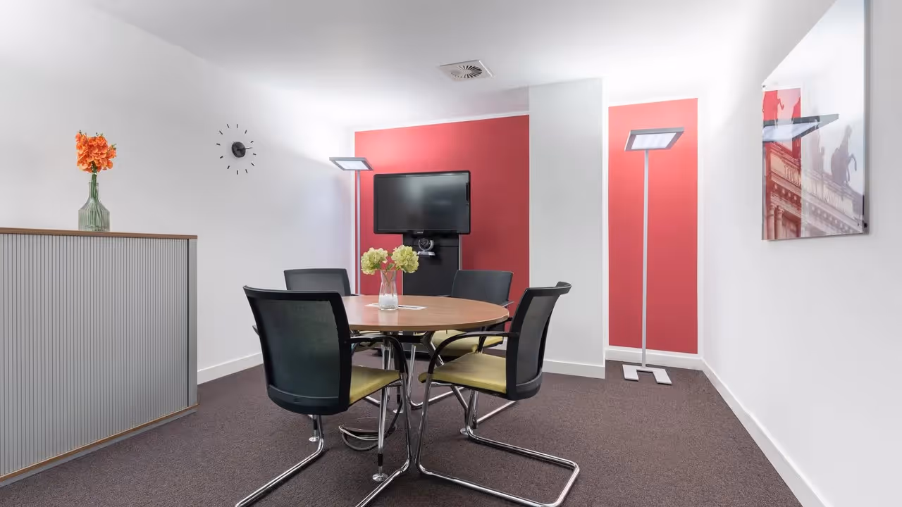 Runder Tisch mit vier Stühlen, Wandmonitor, rote Akzentwand, Stehlampen, Blumenvase, Wanduhr, Kunstbild.