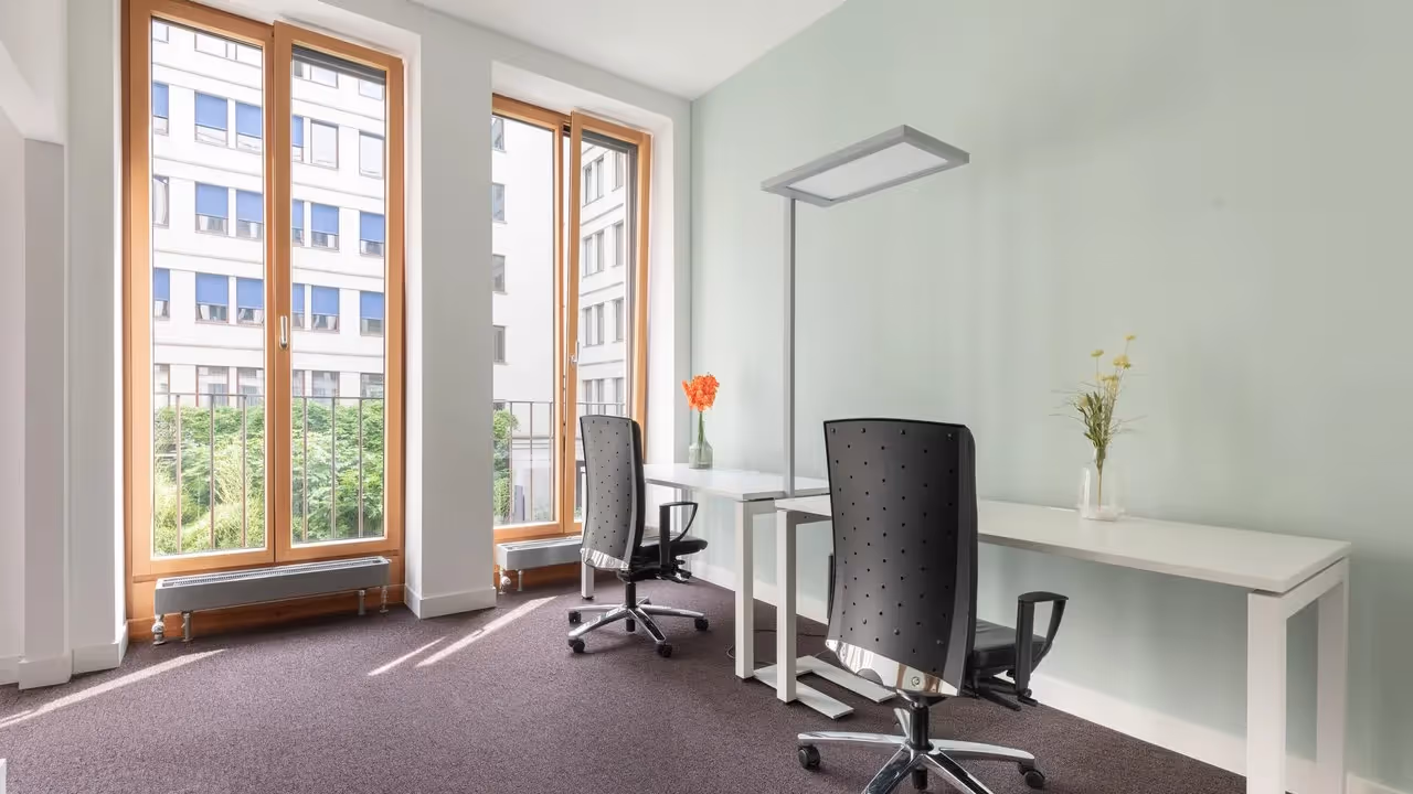 Zwei Schreibtische mit schwarzen Bürostühlen, große Fenster, helle Wände, Blumenvasen, moderne Stehlampe.