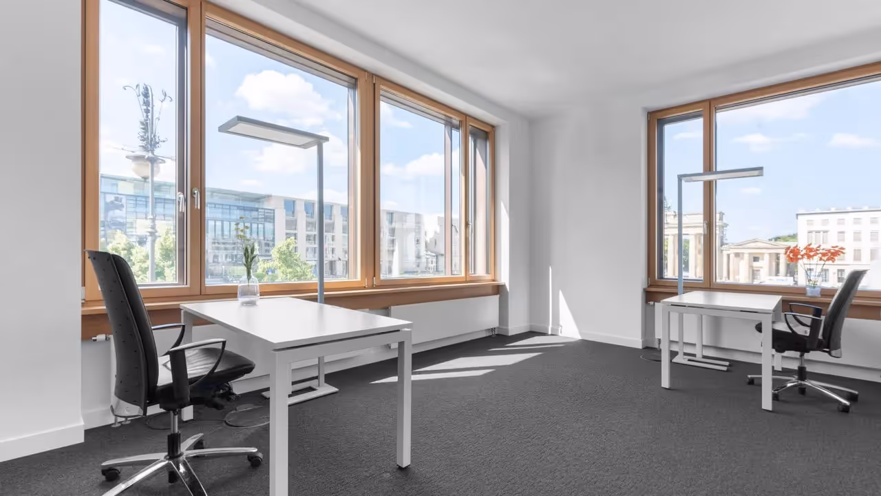 Helles Büro mit zwei Schreibtischen, schwarzen Bürostühlen, großen Fenstern und Blick auf historische Gebäude.
