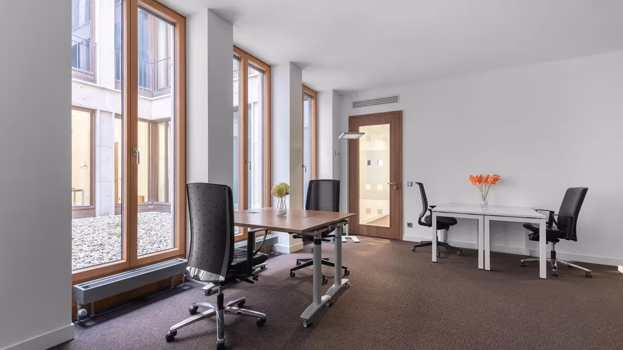 Helles Büro mit großen Fenstern, zwei Schreibtischen, schwarzen Stühlen, Teppichboden und weißen Wänden.