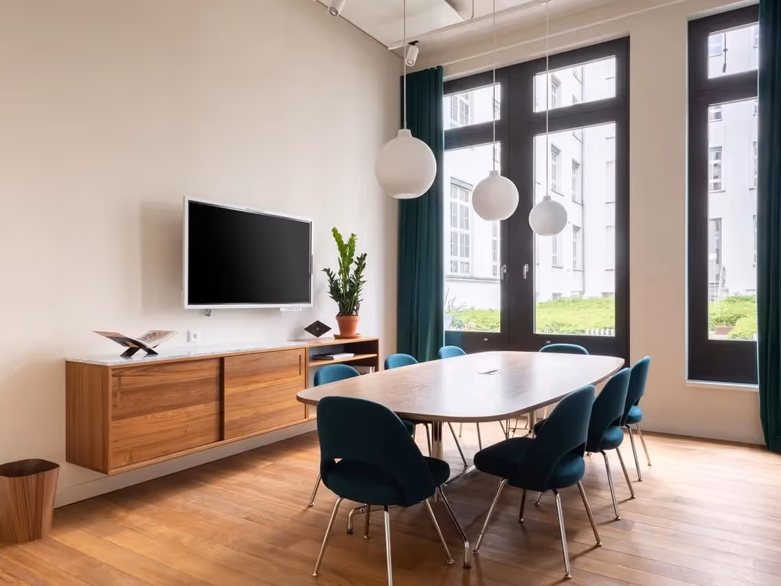 Heller Konferenzraum mit Holztisch, blauen Stühlen, Wandbildschirm, Hängelampen und großen Fenstern.