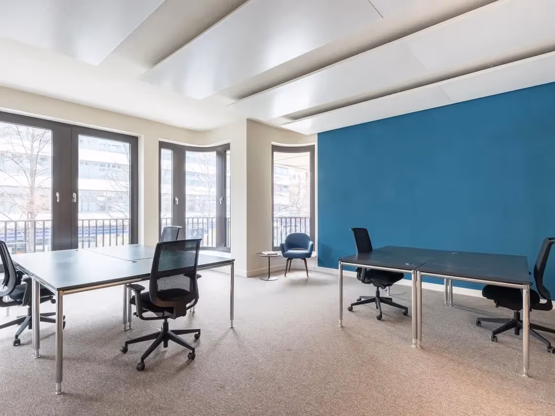 Heller Raum mit vier Schreibtischen, schwarzen Bürostühlen, blauer Wand und großen Fenstern mit Blick nach draußen.