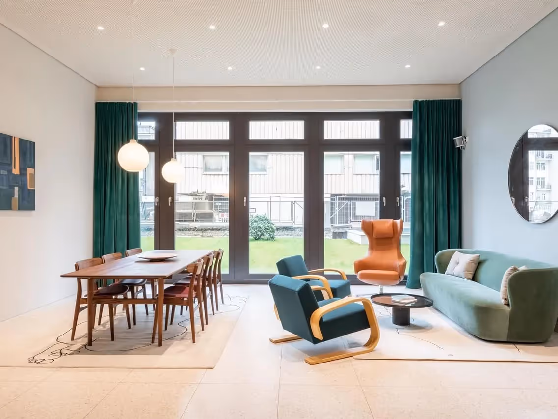 Essbereich mit Holztisch und Stühlen, daneben Sitzecke mit Sofa, Sesseln, rundem Tisch, großen Fenstern und grünen Vorhängen.
