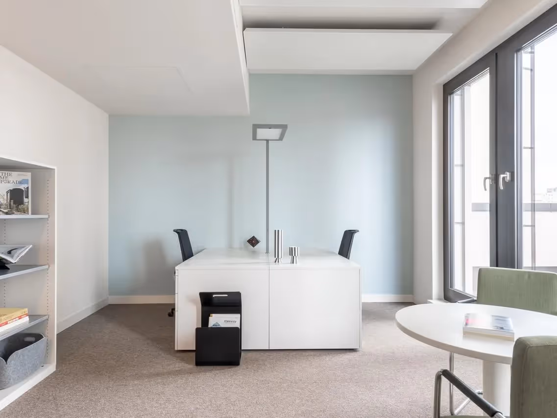 Helles Büro mit zwei Schreibtischen, Stühlen, Regalen, runden Tisch, großen Fenstern und Teppichboden.