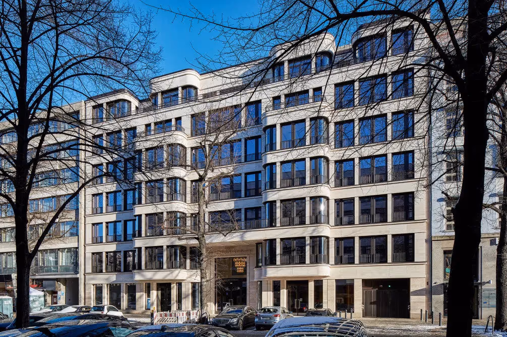 Mehrstöckiges Gebäude mit großen, quadratischen Fenstern, helle Fassade, Bäume im Vordergrund, blauer Himmel.