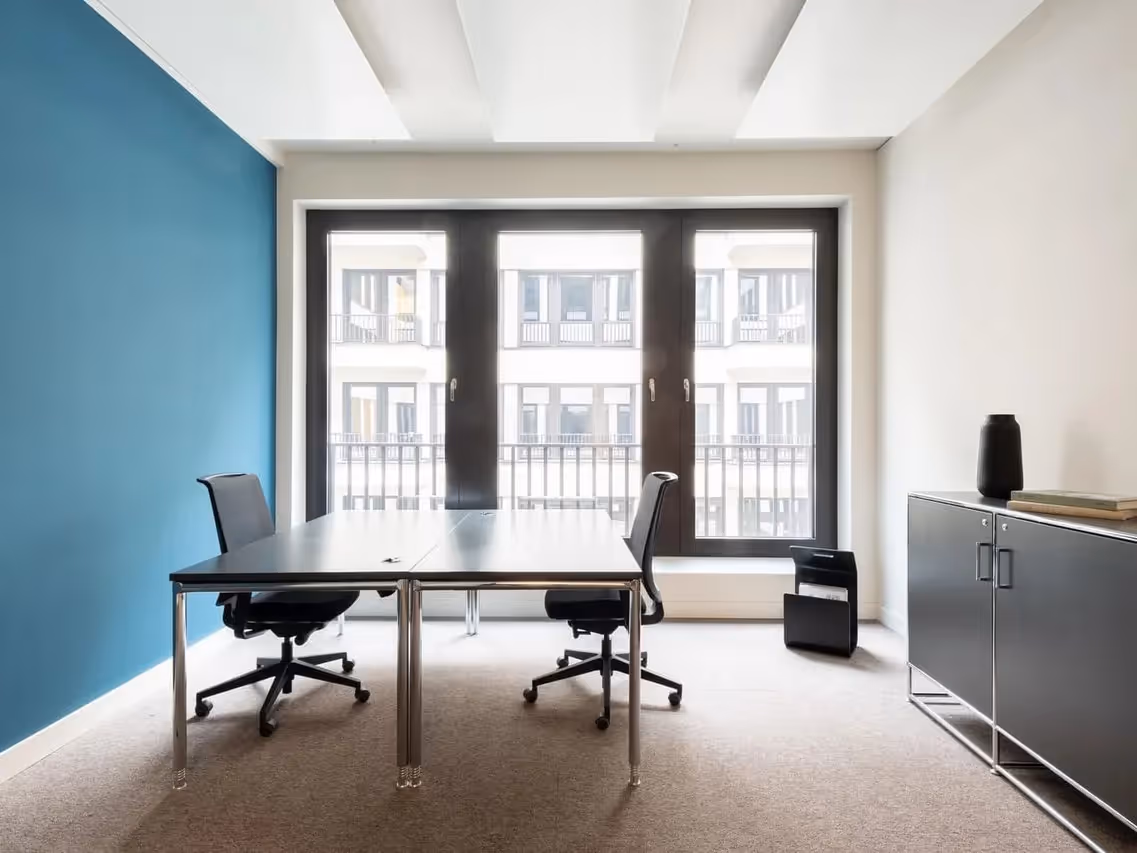 Zwei Bürostühle an einem Tisch, blaue Wand, große Fenster, seitlicher Aktenschrank mit Dekoration.