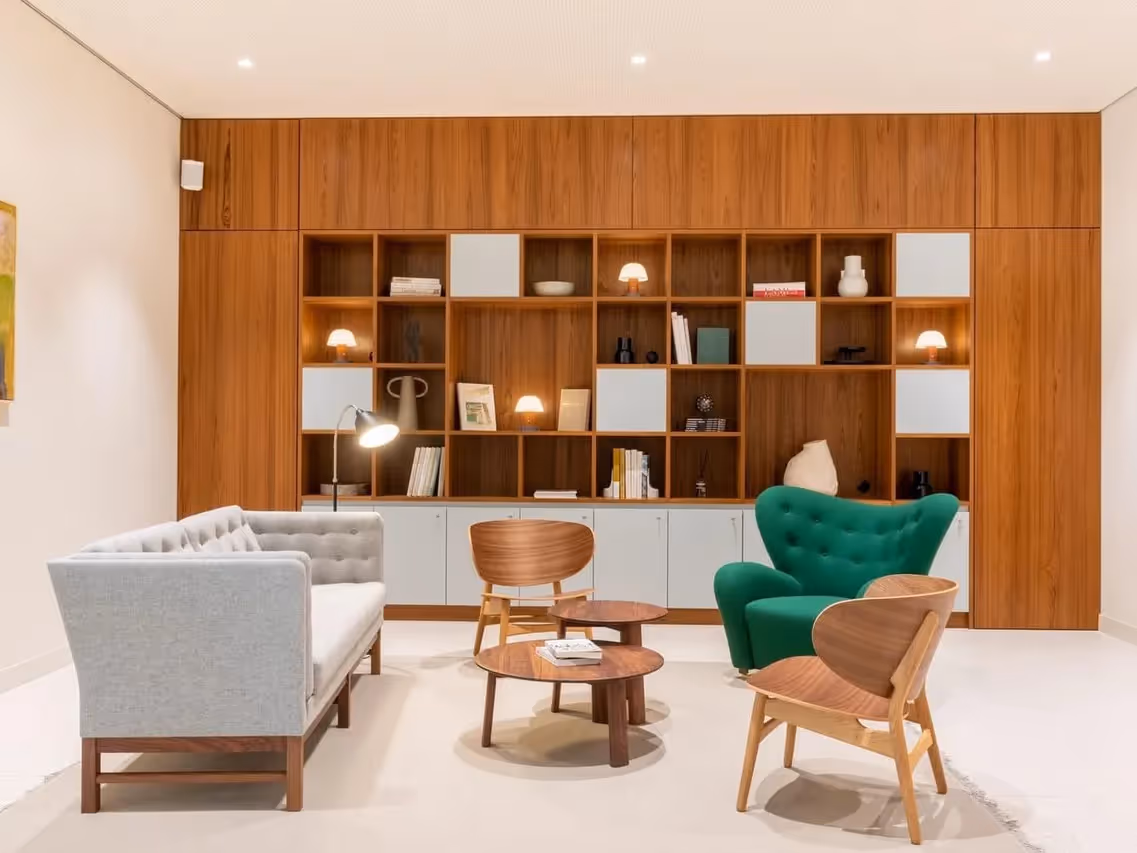 Sitzecke mit grauem Sofa, grünem Sessel, Holztisch und Regalen vor holzgetäfelter Wand mit Beleuchtung.