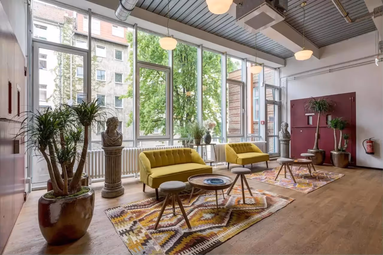 Heller Raum mit gelben Sofas, bunten Teppichen, großen Fenstern, Pflanzen in Töpfen und Büsten auf Sockeln.