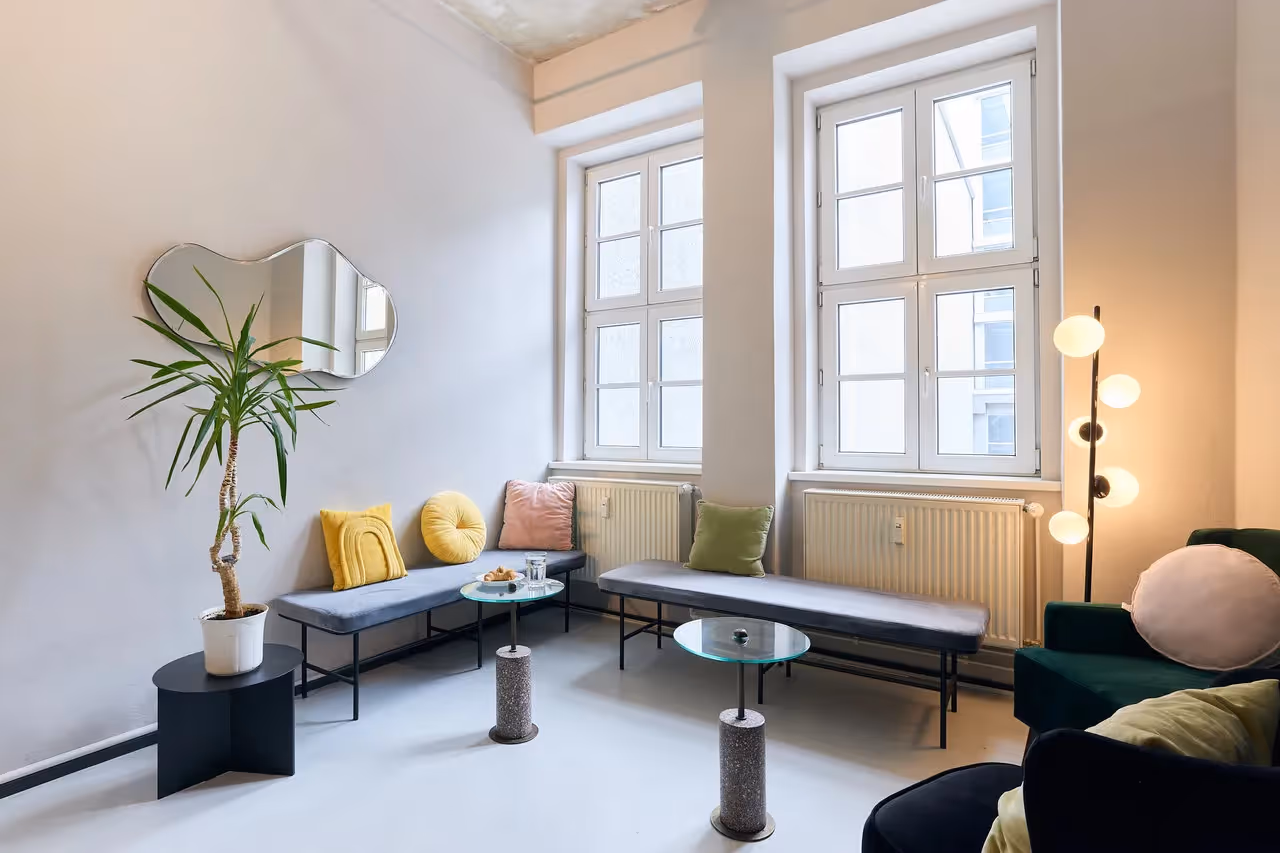 Lounge mit grauen Bänken, bunten Kissen, Glas-Couchtischen, Stehlampe, Pflanze und großen Fenstern.