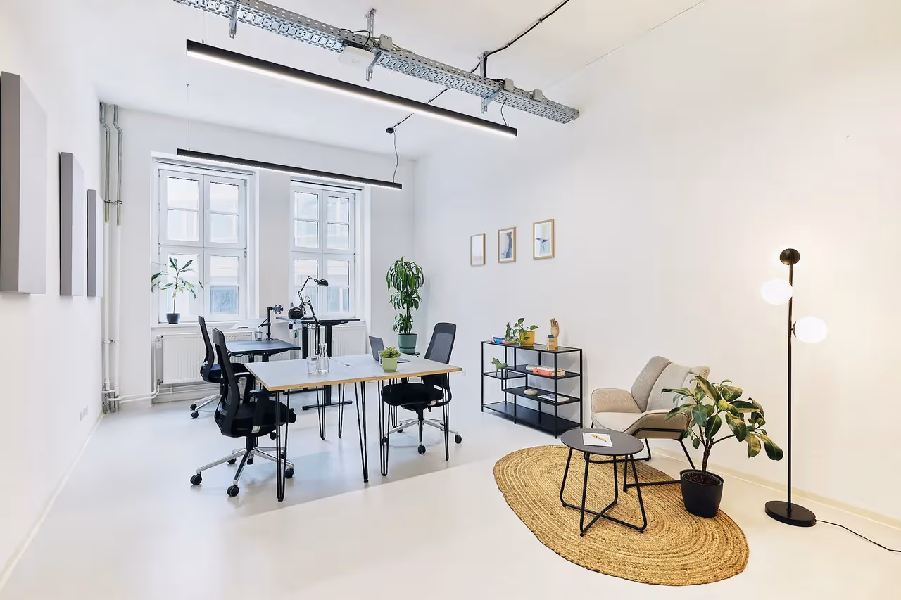 Heller Raum mit Schreibtischen, Stühlen, Pflanzen, Stehlampe, Sofa, Teppich, Regalen und großen Fenstern.