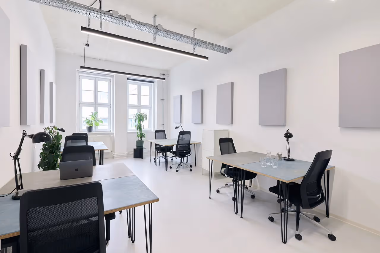 Heller Raum mit Schreibtischen, schwarzen Stühlen, Pflanzen, Lampen und großen Fenstern, minimalistisches Design.