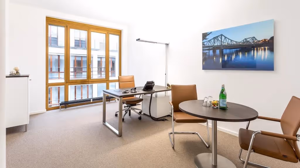 Helles Büro mit großem Fenster, Schreibtisch, zwei braunen Stühlen, rundem Tisch, Bild an der Wand und Teppichboden.