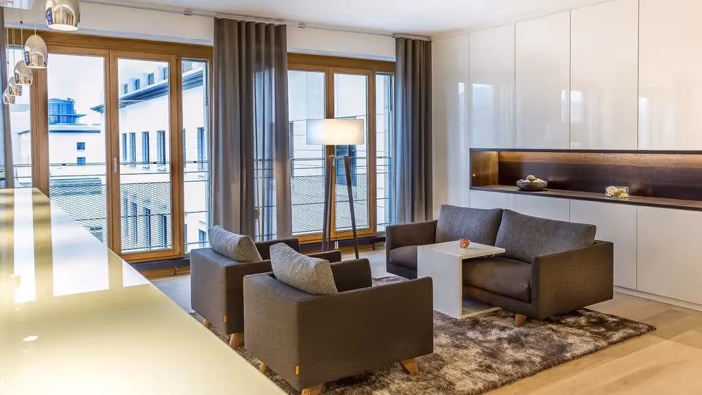 Zwei graue Sessel und Sofa um weißen Tisch, große Fenster, Holzboden, Deckenleuchten, helle Wände, Teppich.