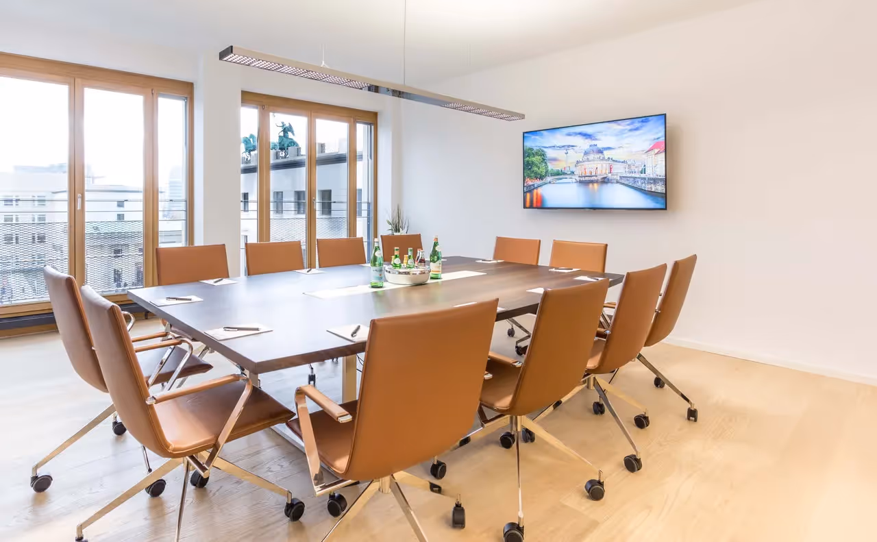 Konferenzraum mit Holztisch, braunen Drehstühlen, großem Fenster, Wandbildschirm und hängender Deckenleuchte.