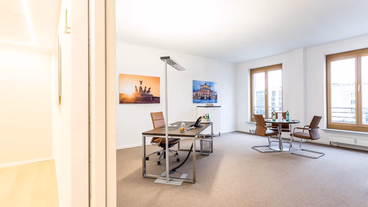 Helles Büro mit braunem Schreibtisch, Ledersessel, Rundtisch, großen Fenstern und Wandbildern.