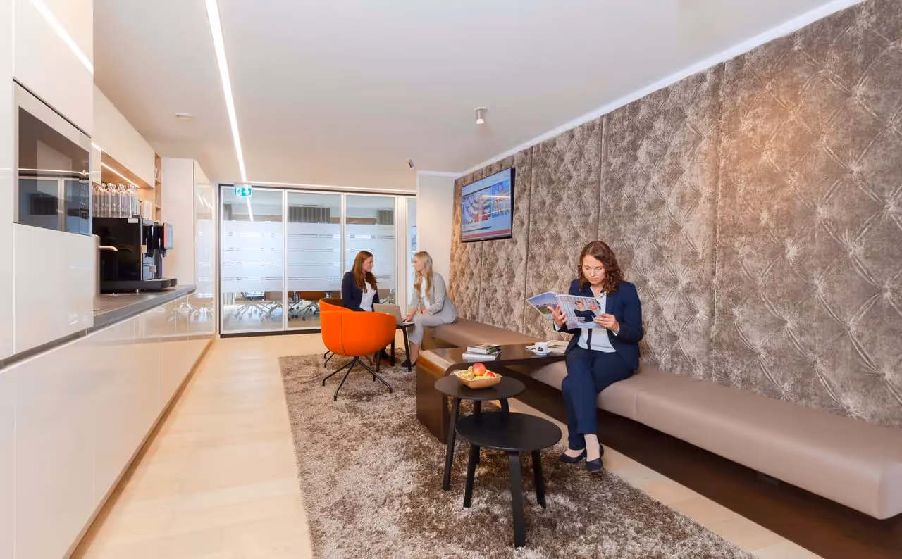 Drei Personen in eleganter Lounge mit Teppich, Ledersitzbank, orangenem Sessel, Couchtischen und Kaffeemaschine.