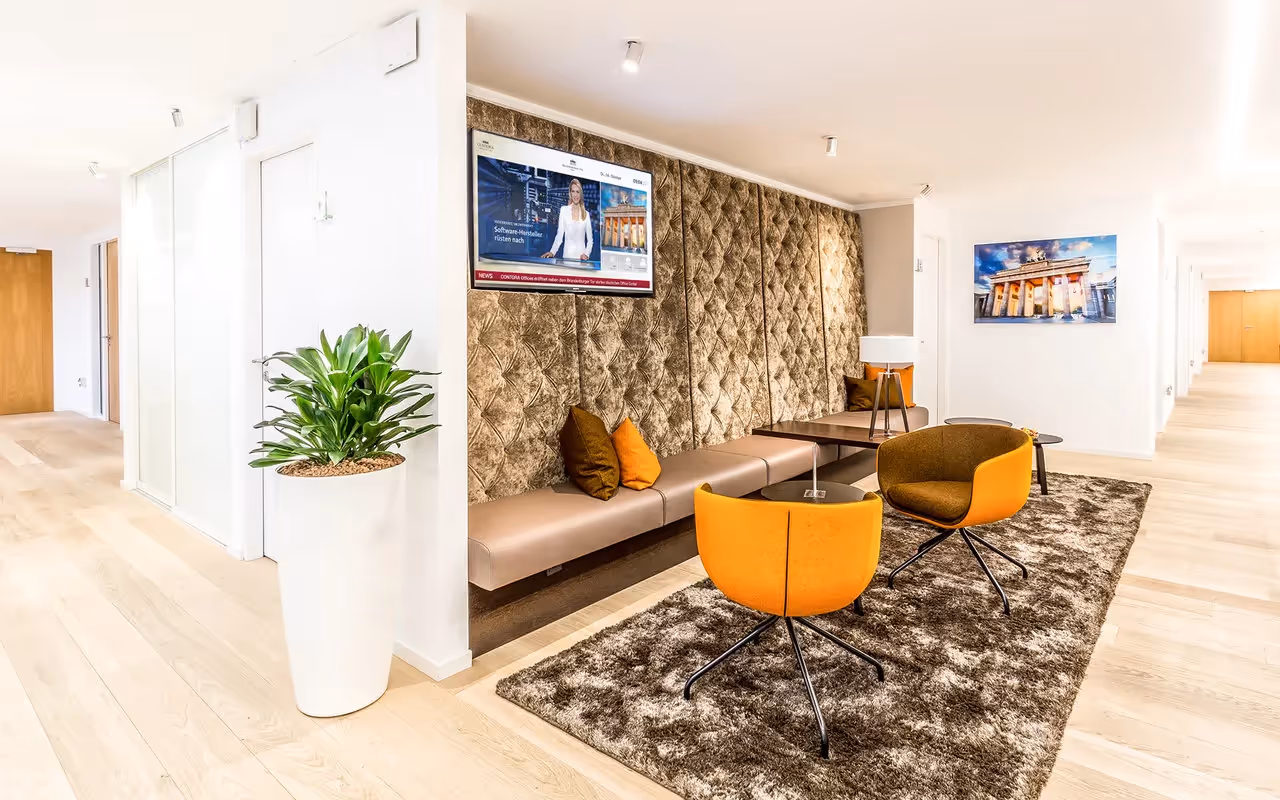 Loungebereich mit brauner Wandpolsterung, orange Stühlen, braunem Teppich und großem Pflanztopf auf Holzboden.