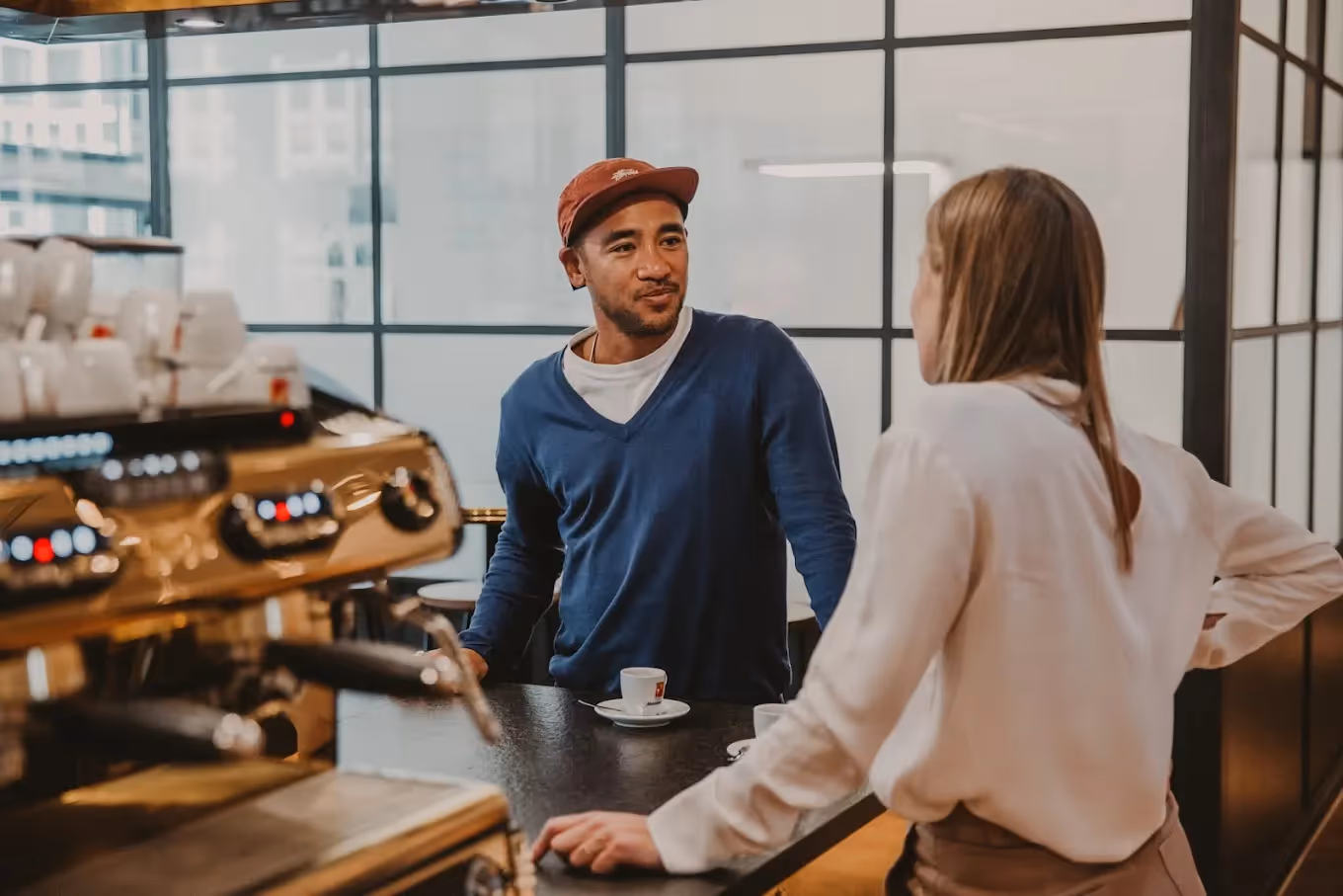 Zwei Personen unterhalten sich an einer Kaffeebar mit Espressomaschine, im Hintergrund große Fenster.