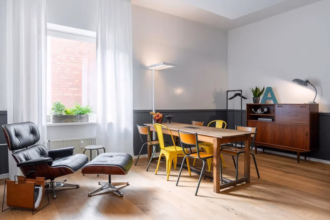Heller Raum mit Esstisch, gelben Stühlen, Sessel, Sideboard, Stehlampe, Pflanzen, Holzfußboden und großen Fenstern.