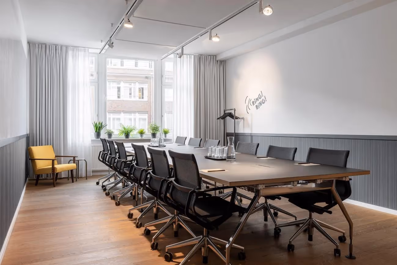 Konferenzraum mit großem Tisch, schwarzen Bürostühlen, gelbem Sessel, Pflanzen am Fenster, helle Vorhänge.