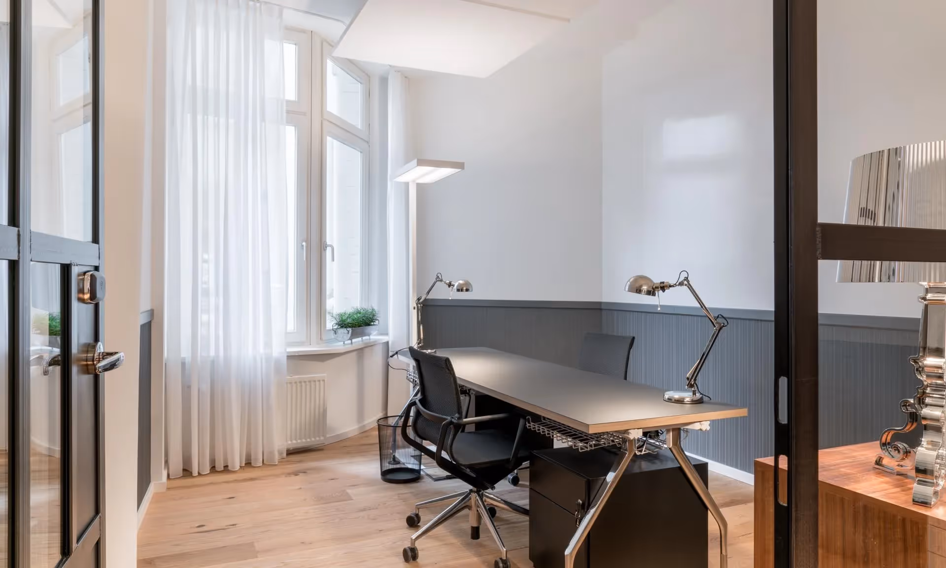 Helles Büro mit großem Fenster, Holzboden, Schreibtisch, zwei Stühlen, Metalllampen und Pflanzen auf Fensterbank.