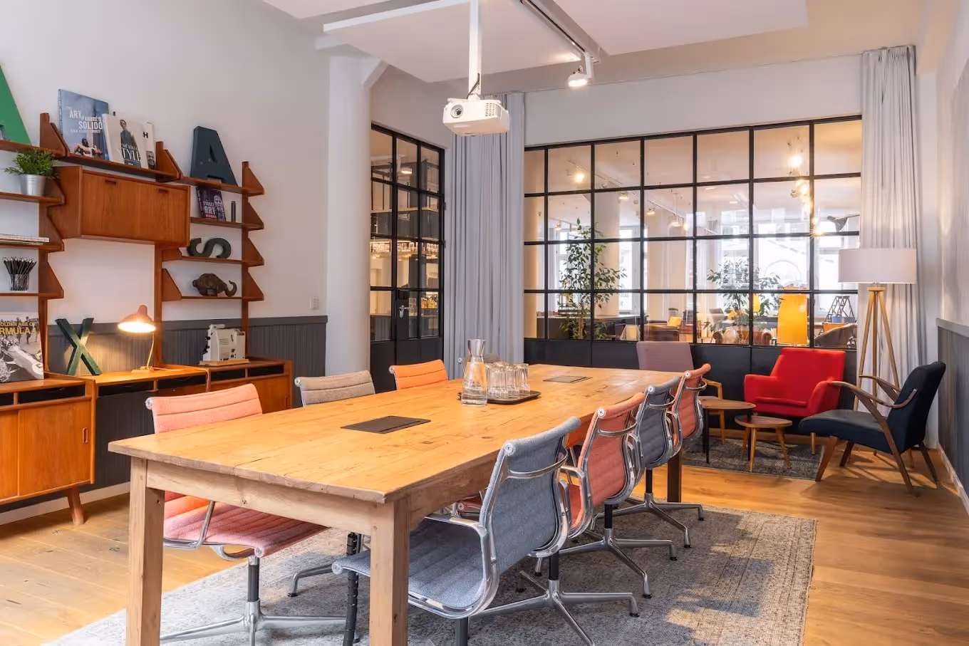 Großer Holztisch mit bunten Stühlen, Retro-Regal, Projektor, Glaswand, rote und blaue Sessel, Stehlampe.