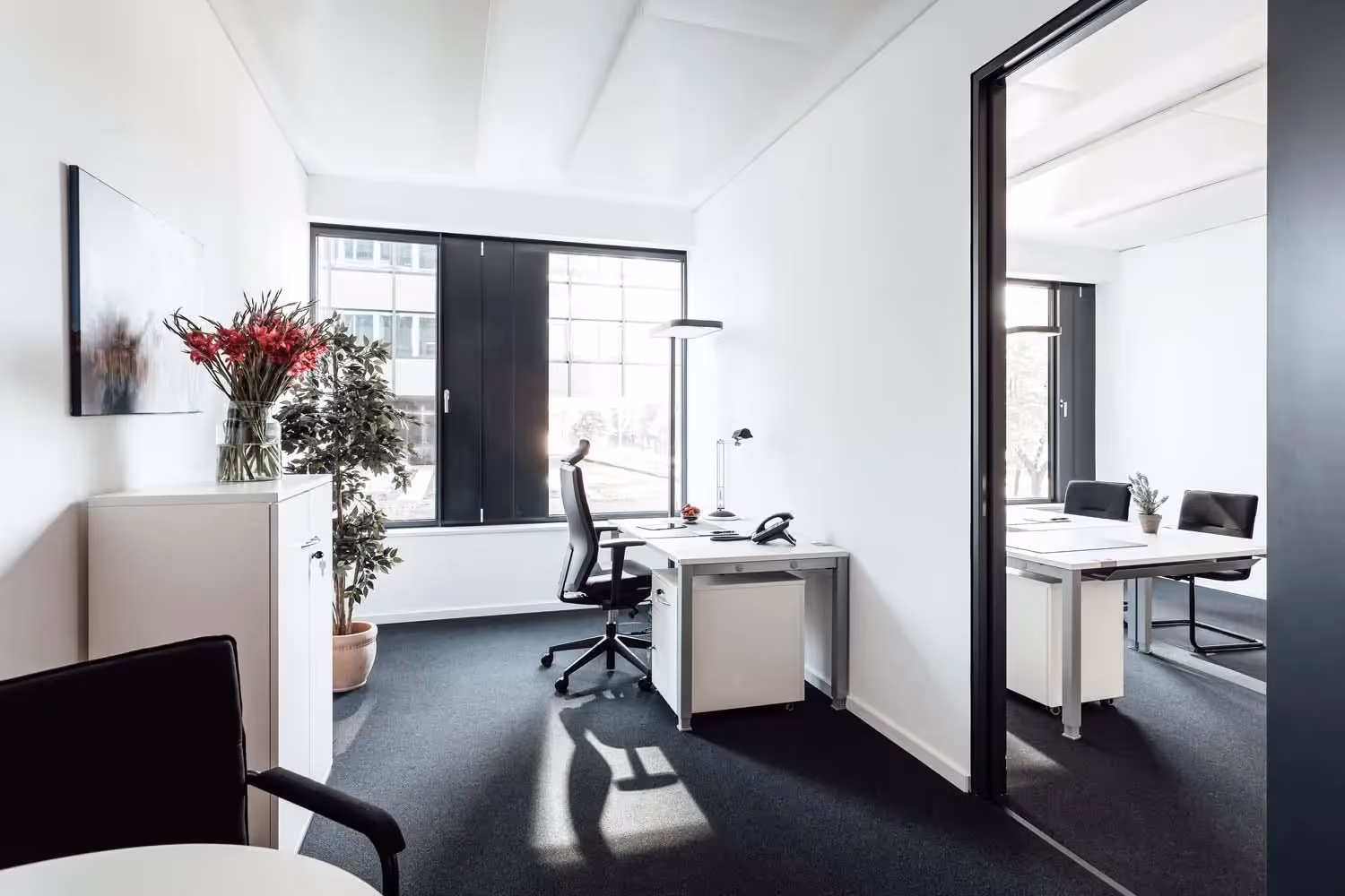 Helles Büro mit Schreibtisch, Bürostuhl, Pflanzen, großem Fenster und angrenzendem Konferenzraum mit Tisch und Stühlen.