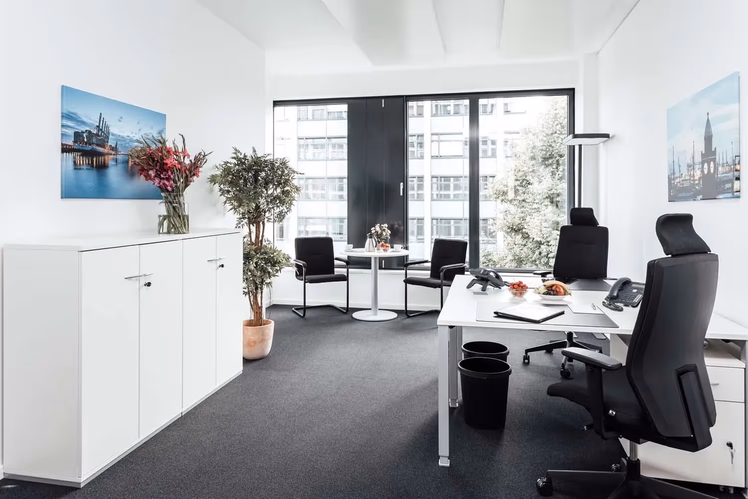 Heller Raum mit Schreibtisch, zwei Stühlen, Pflanzen, großem Fenster und Schränken, dekoriert mit Hafenbildern.
