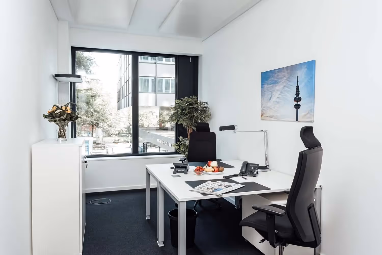 Helles Büro mit zwei Schreibtischen, schwarzen Bürostühlen, Fenster, Pflanzen, Lampe, Bild an der Wand, Obst auf Tisch.