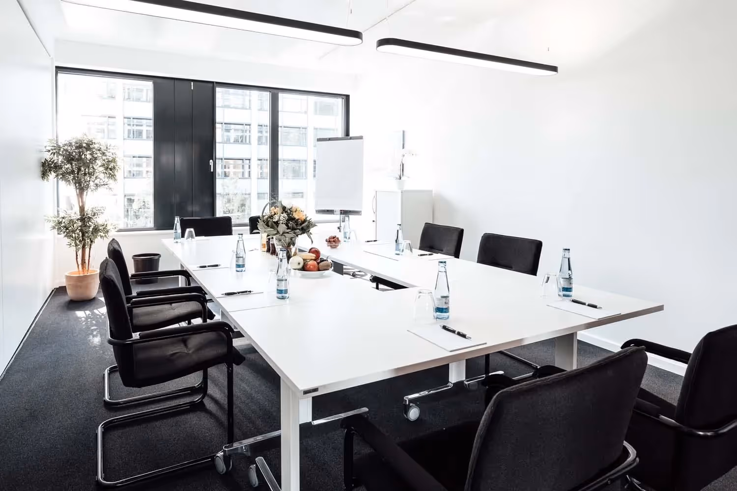 Heller Besprechungsraum mit großem Tisch, schwarzen Stühlen, Pflanzen, Fenstern, Flipchart und Beleuchtung.