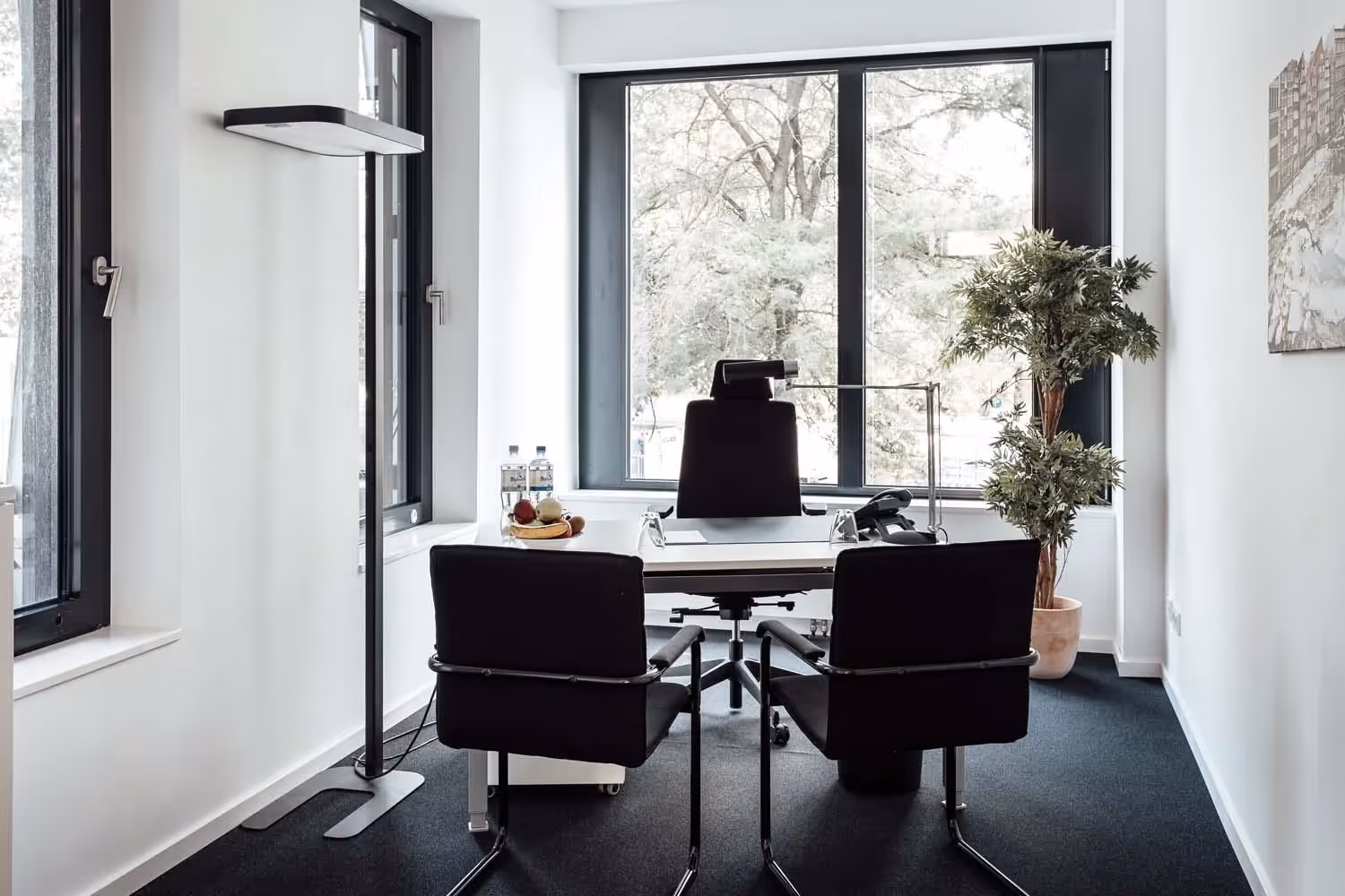 Helles Büro mit großem Fenster, Schreibtisch, zwei Stühlen, Stehlampe, Pflanze und Obstschale.