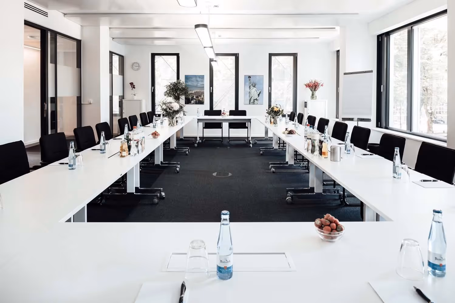 Konferenzraum mit U-förmiger Tischanordnung, schwarzen Stühlen, Wasserflaschen und Blumen, helle Fensterfront.