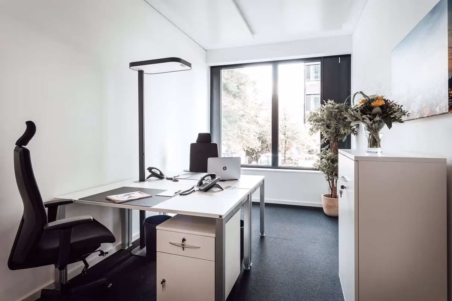 Helles Büro mit großem Fenster, zwei Schreibtischen, Bürostühlen, Pflanzen und einem Schrank mit Blumenstrauß.