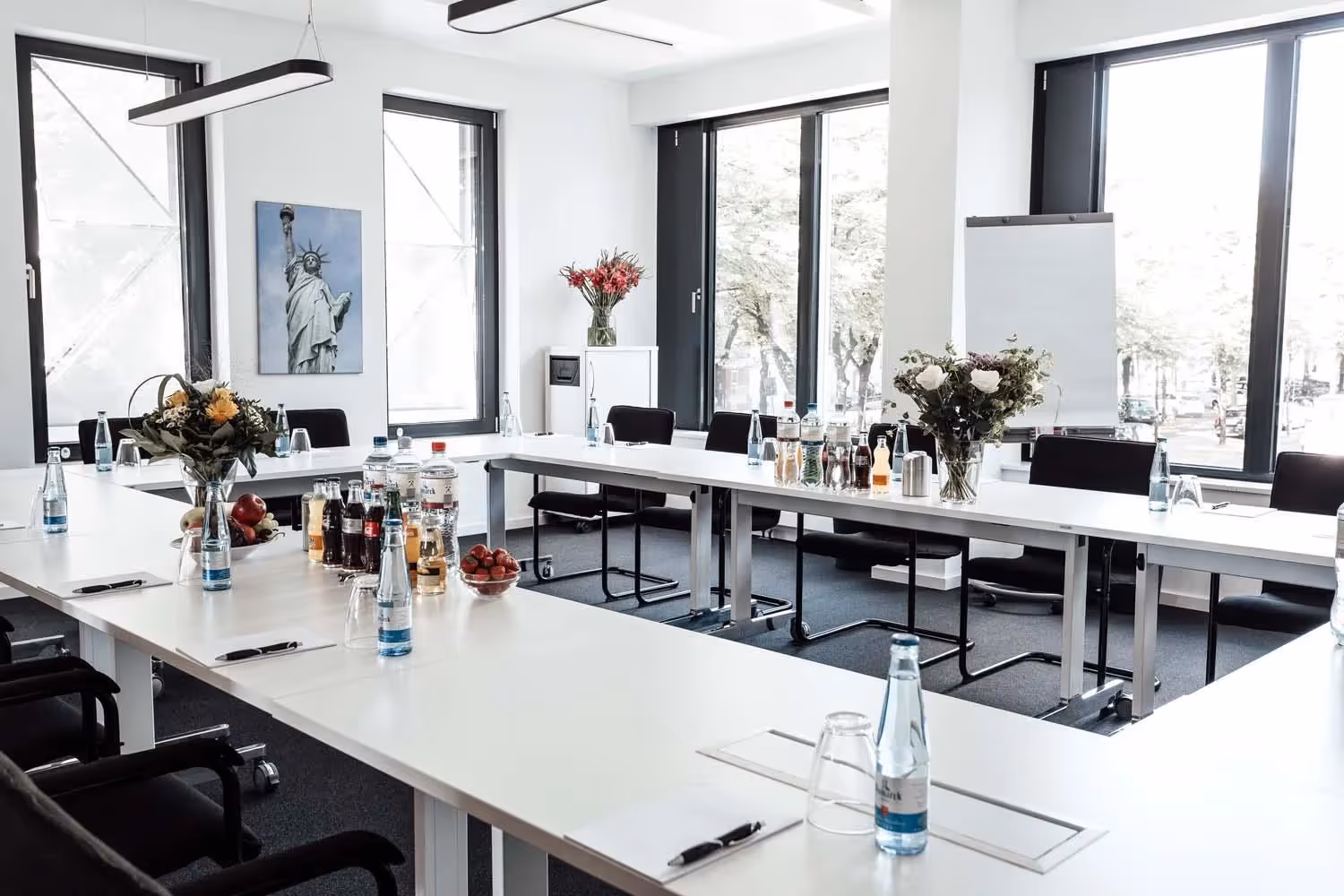 Heller Konferenzraum mit U-förmigem Tisch, Blumen, Getränken, großen Fenstern und einem Bild der Freiheitsstatue.