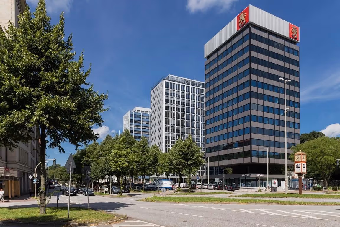 Mehrere moderne Bürogebäude mit Glasfassaden, umgeben von Bäumen und parkenden Autos, unter blauem Himmel.