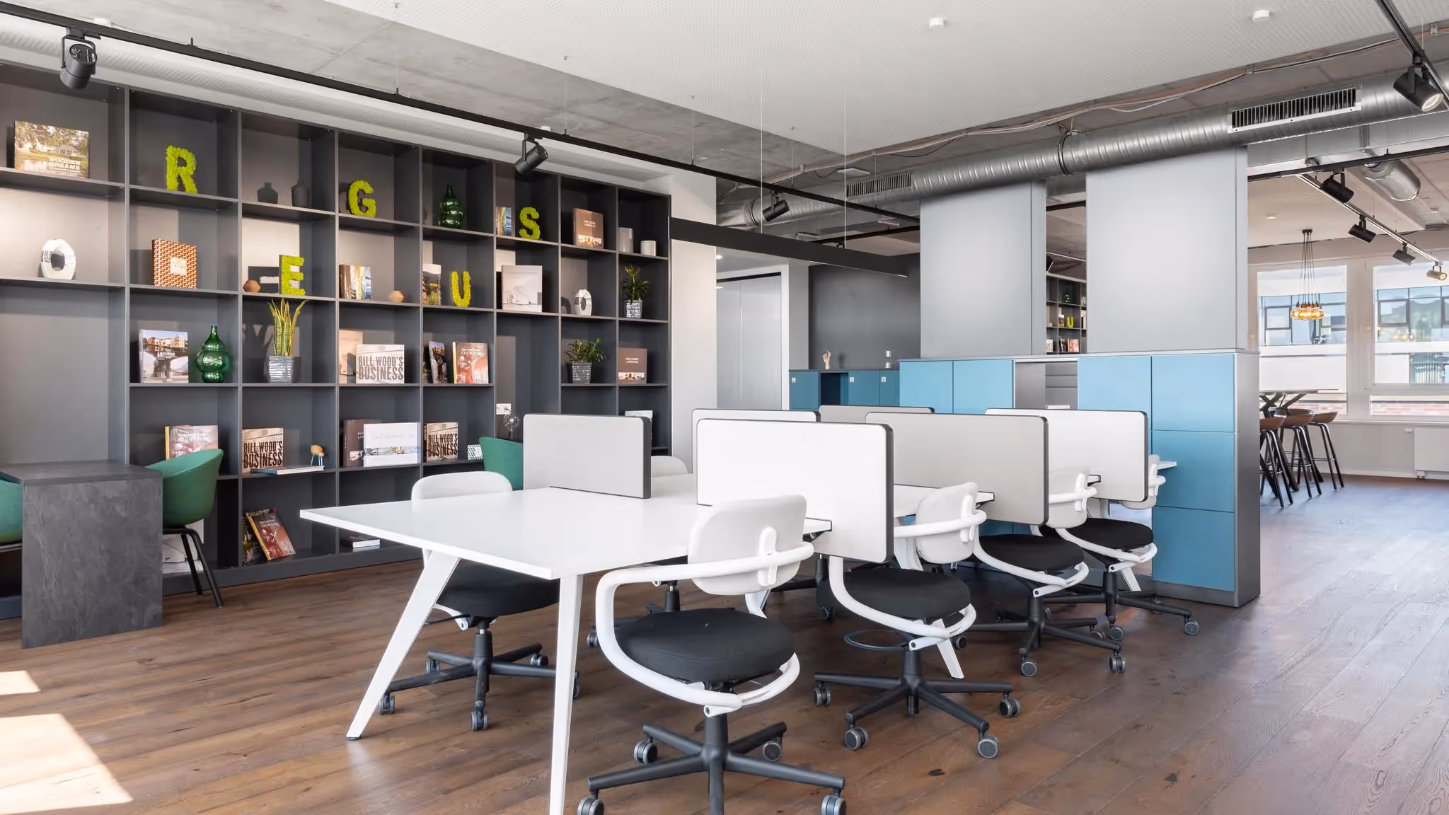 Helles Büro mit weißen Schreibtischen, schwarzen Drehstühlen, Regalen, Holzfußboden und blauen Schränken.
