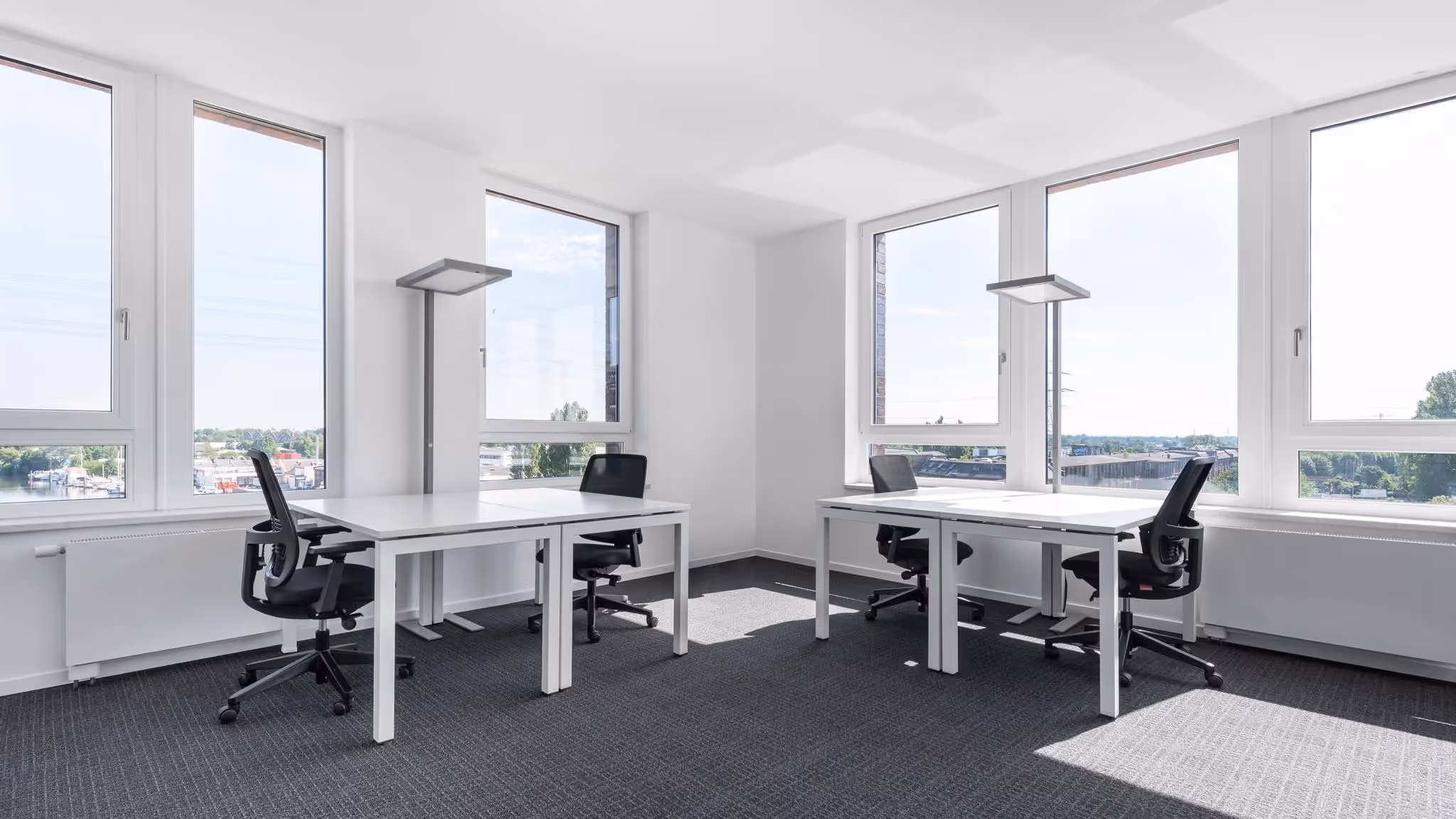 Helles Büro mit drei weißen Schreibtischen, schwarzen Bürostühlen, großen Fenstern und grauem Teppichboden.