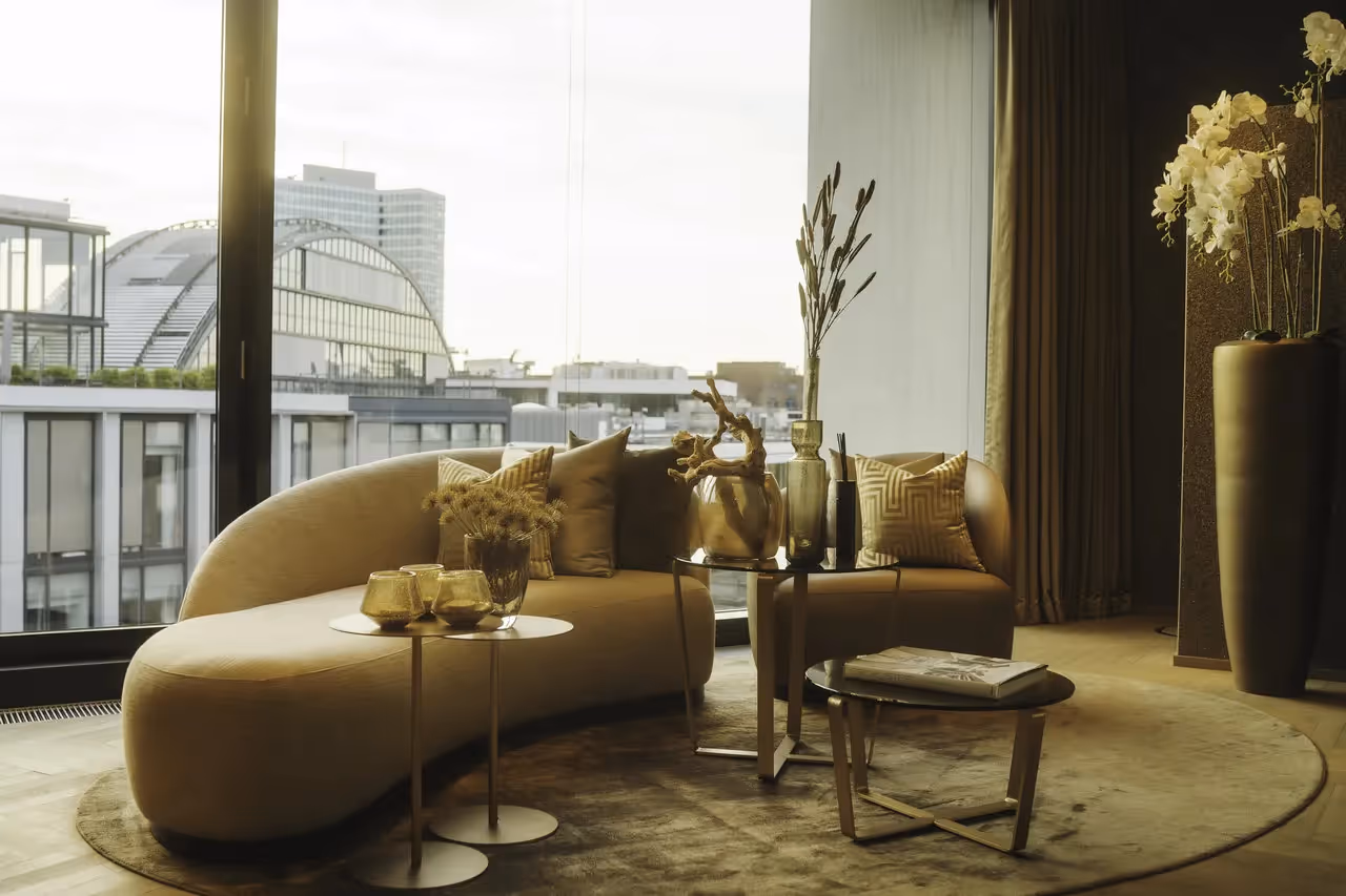 Gebogener beiger Sofa mit Kissen, runde Tische, große Fensterfront, Dekovasen, Teppich, Blick auf Stadtgebäude.