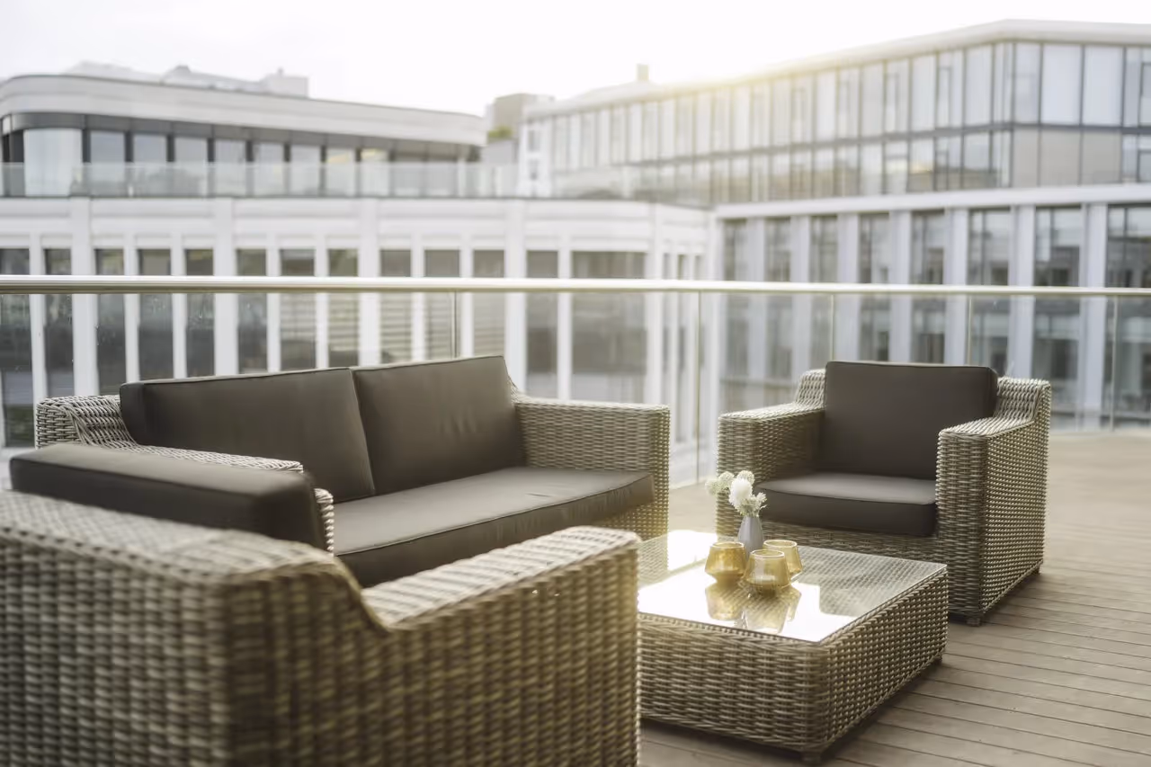 Geflochtene Sitzgruppe mit Glastisch auf einer Terrasse, im Hintergrund moderne Bürogebäude.