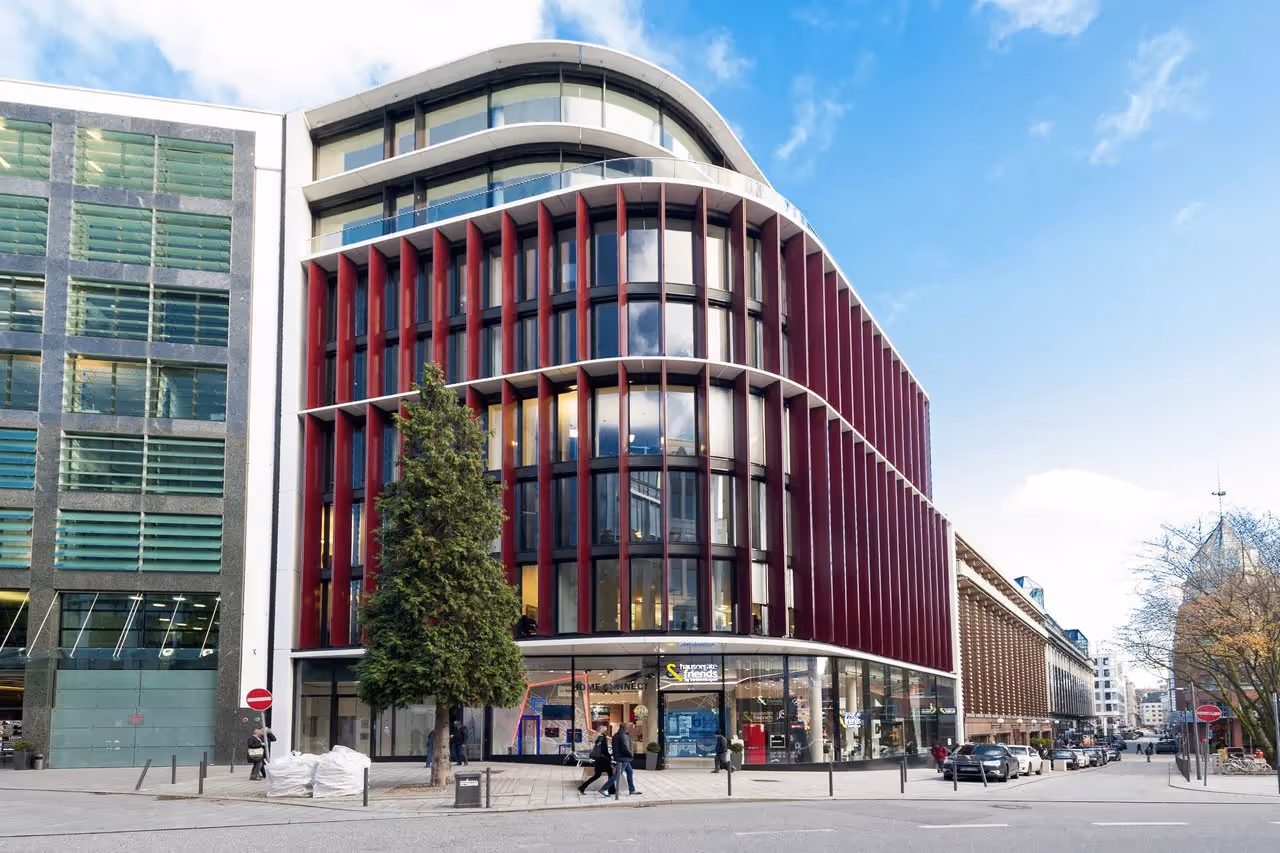 Gebäude mit roter Fassade, Glasfronten, geschwungenen Linien, Fußgänger und Bäume im Vordergrund, blauer Himmel.