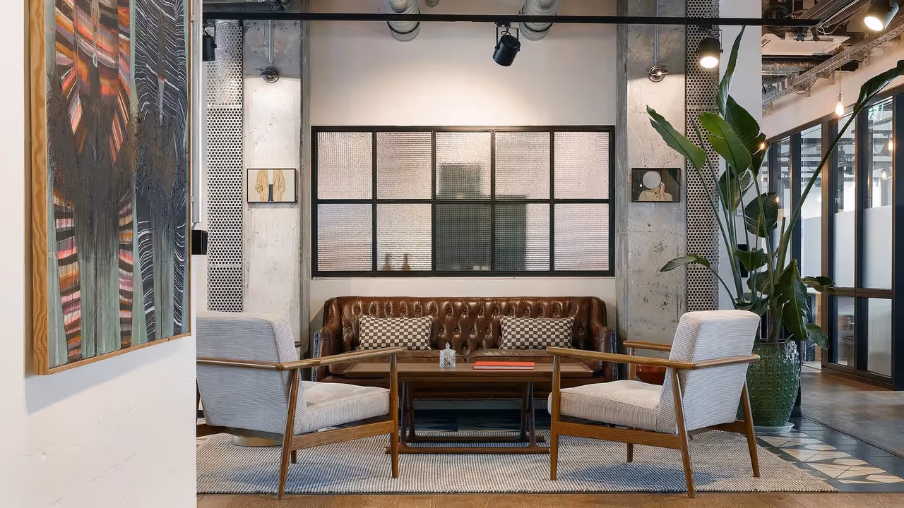 Braune Ledercouch mit karierten Kissen, zwei graue Sessel, großer Pflanzenkübel, Wandkunst, Glaswand im Hintergrund.
