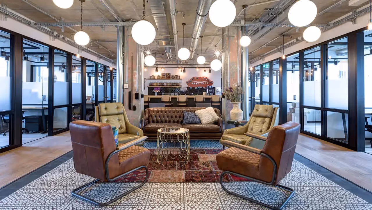 Lounge mit Ledersesseln, Teppich, runden Tischen, Bar im Hintergrund, Glaswände, Hängelampen, industrieller Stil.